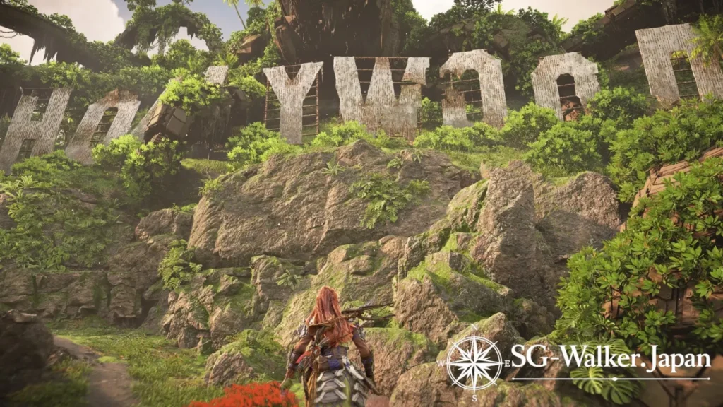 『Horizon Forbidden West: Burning Shores』で、植物に覆われた山肌に朽ちたハリウッドサインを見上げるアーロイ